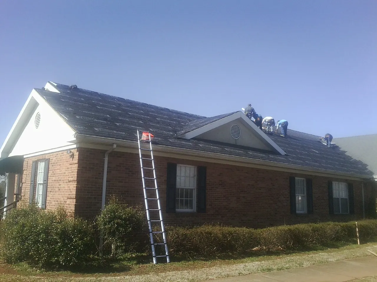 Crew tearing shingles off a brick ranch roof with a ladder set — Emergency Roof Repair work by our crew in Goodspring