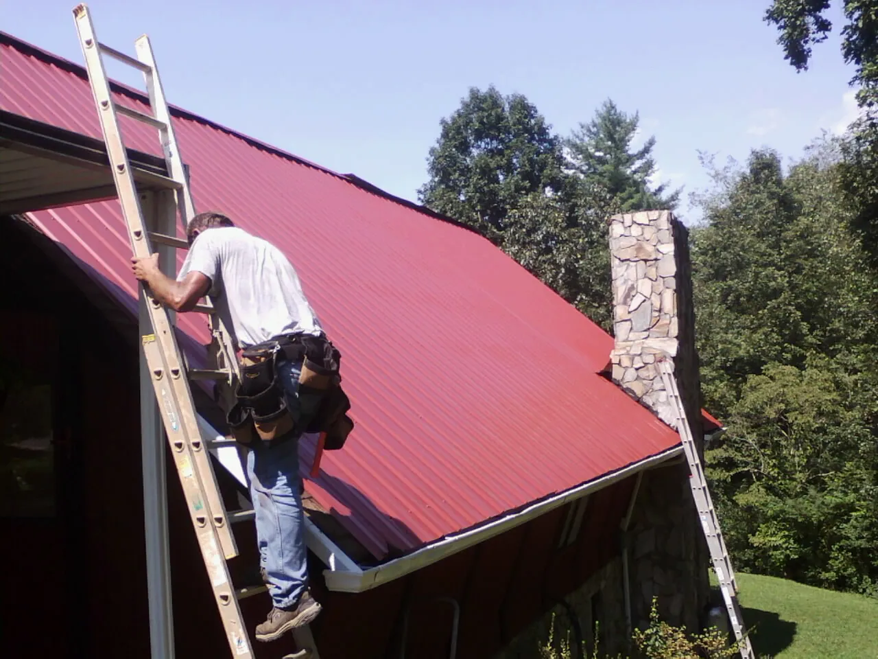 Roofer on a ladder beside a red metal roof and stone chimney — Soffit & Fascia Repair by Roofing Brothers in Goodspring